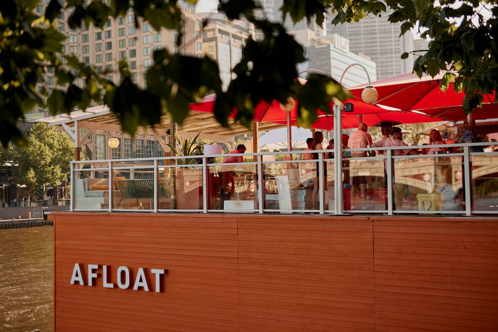 Timber panelling on the side of Arbory's Afloat, with the text ''Afloat" written in white font. On the vessel are umbrellas and people sitting on deck chairs.