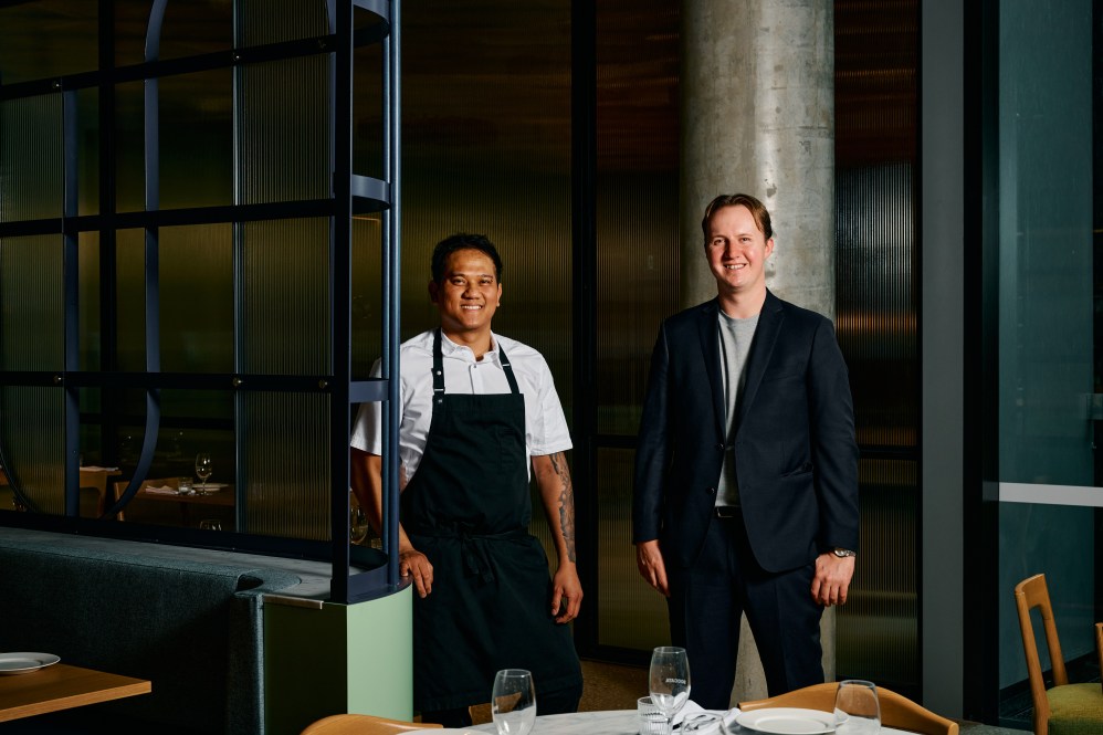 Christian Corpuz in a white shirt and apron and Dylan Wisnesky in a black suit and grey shirt stand in the low-lit interior of Boccata.