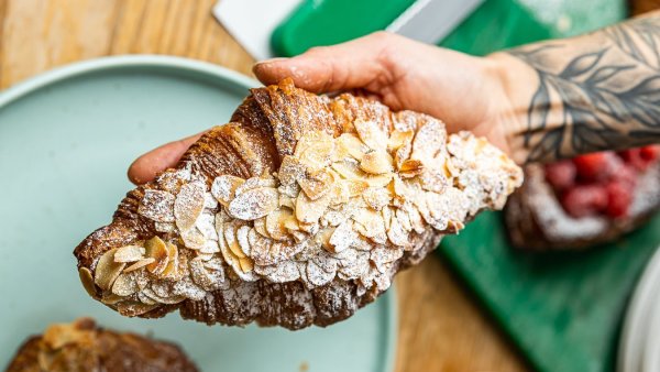 A person with a tattooed arm holds an Oh My Days almond croissant in one hand.