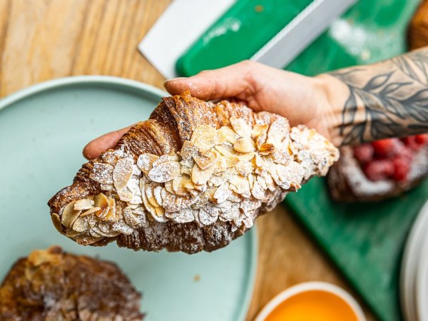 A person with a tattooed arm holds an Oh My Days almond croissant in one hand.