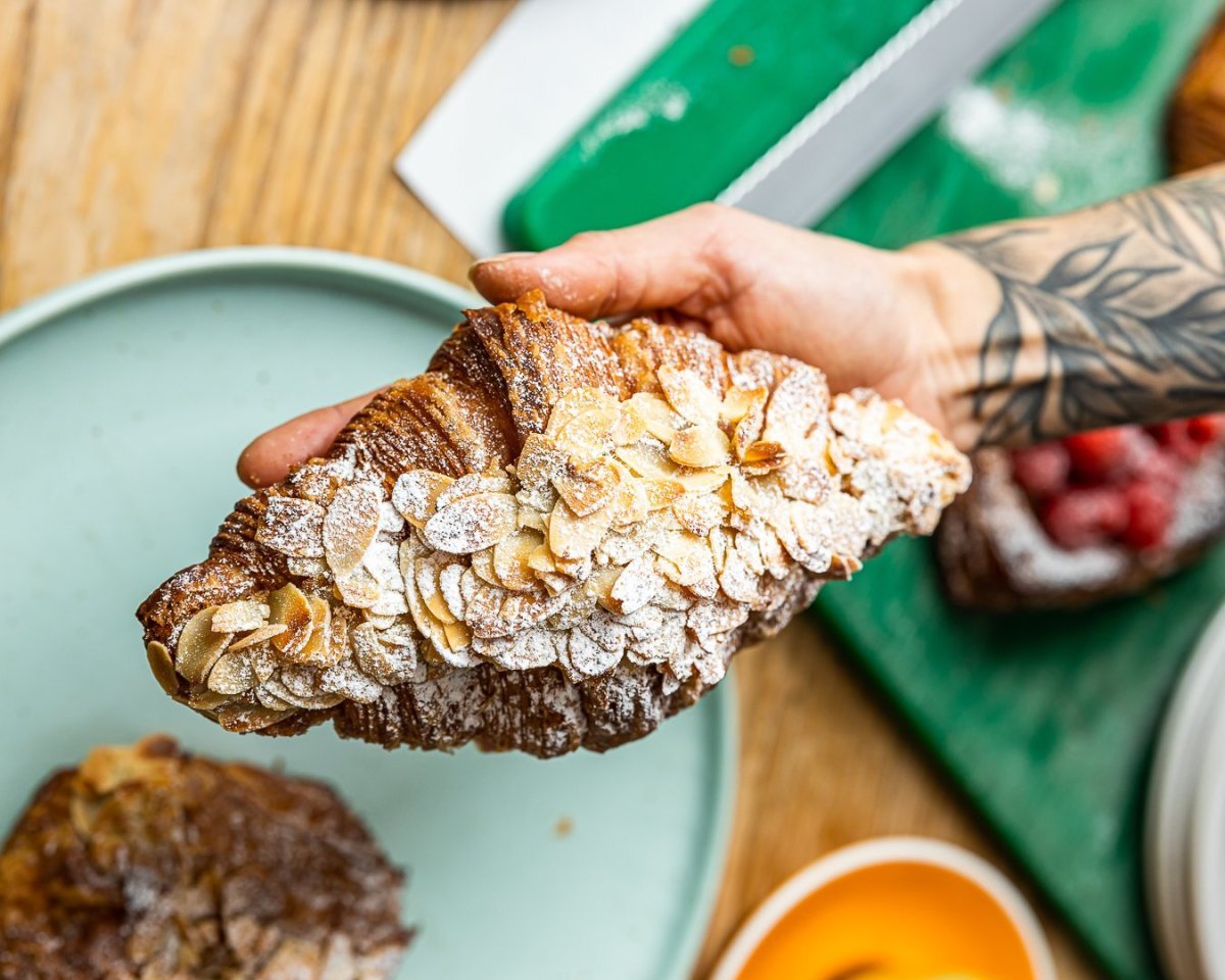 A person with a tattooed arm holds an Oh My Days almond croissant in one hand.