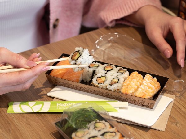 A woman uses wooden chopsticks to eat sushi from a BioPak sushi tray.