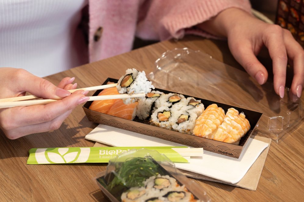 A woman uses wooden chopsticks to eat sushi from a BioPak sushi tray.