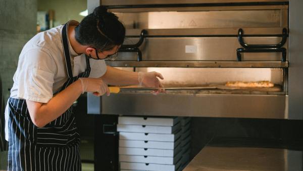 A hospitality worker puts pizza into an oven.