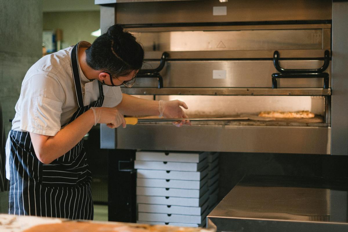 A hospitality worker puts pizza into an oven.