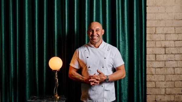 Shane Delia wears chef's whites and stands in front of a green curtain.