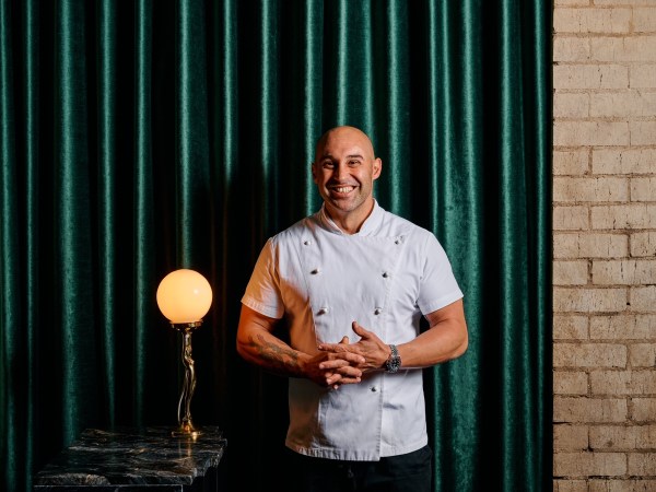 Shane Delia wears chef's whites and stands in front of a green curtain.