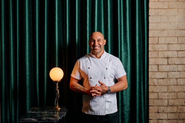 Shane Delia wears chef's whites and stands in front of a green curtain.